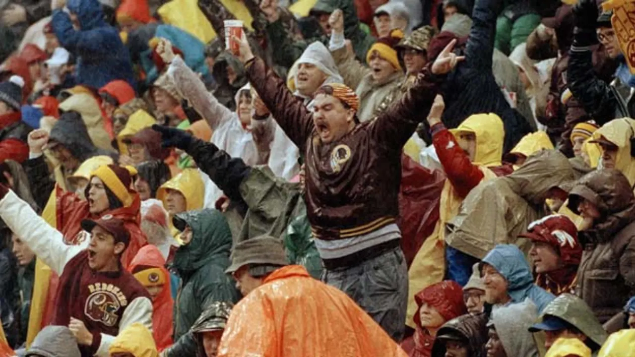 Washington Redskins fans celebrate in the stands at RFK Stadium during the rainy 1991 NFC Divisional Playoff game against the Atlanta Falcons, many wearing team apparel and ponchos with arms raised in excitement